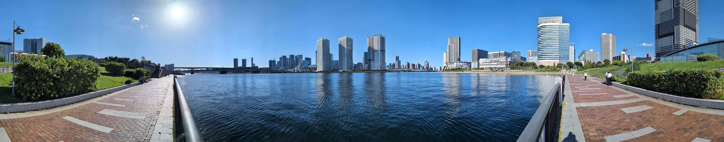 A wide panorama of the Toyosu waterfront from a brick promenade, with towers across the bay under a blue sky; a general photograph of the area, not of the museum