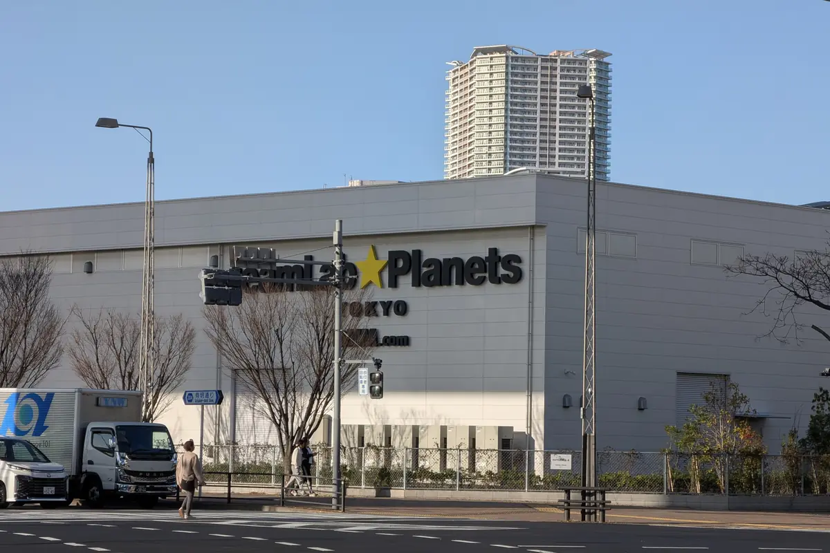 The grey exterior wall of the teamLab Planets building in Toyosu on a clear day, with the museum's name on the facade, bare trees and a pedestrian crossing in front, and a residential tower rising behind