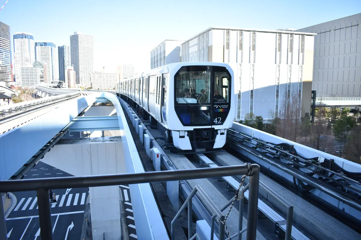 A white Yurikamome train on its elevated guideway above a street junction, with glass towers in the background under a pale blue sky