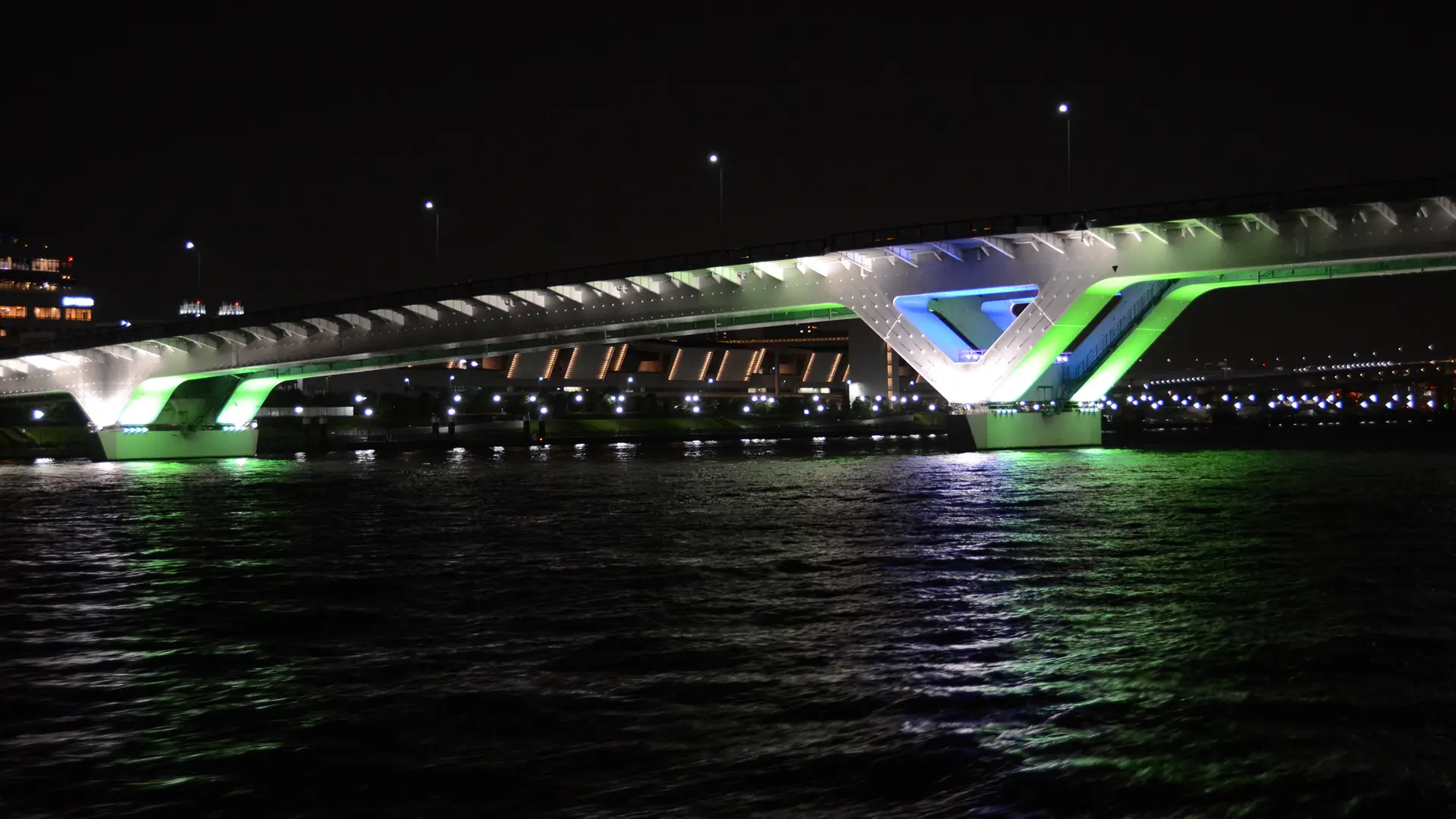 Toyosu Ōhashi bridge lit in green, blue and white above dark water at night, with the lights reflected in the ripples; a general photograph of the Toyosu waterfront, not of teamLab Planets or its artworks